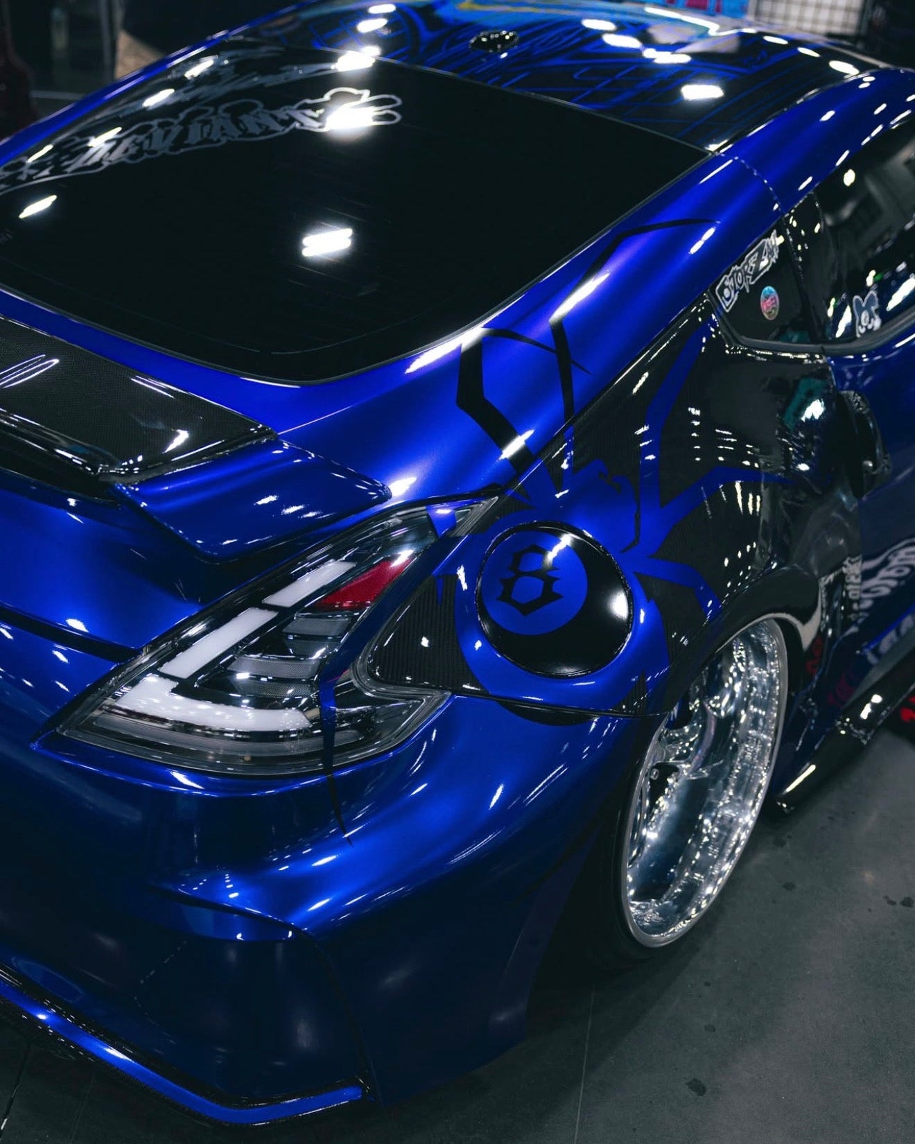 A glossy deep blue custom car with black graphics, custom chrome wheels, and a lowered stance, photographed at an indoor auto show.