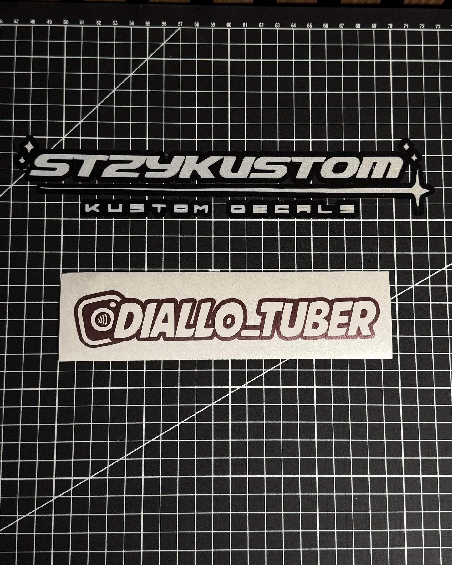 Two stickers on a grid surface, one labeled 'STRYKUSTOM' and the other 'DIALLO_TUBER'.