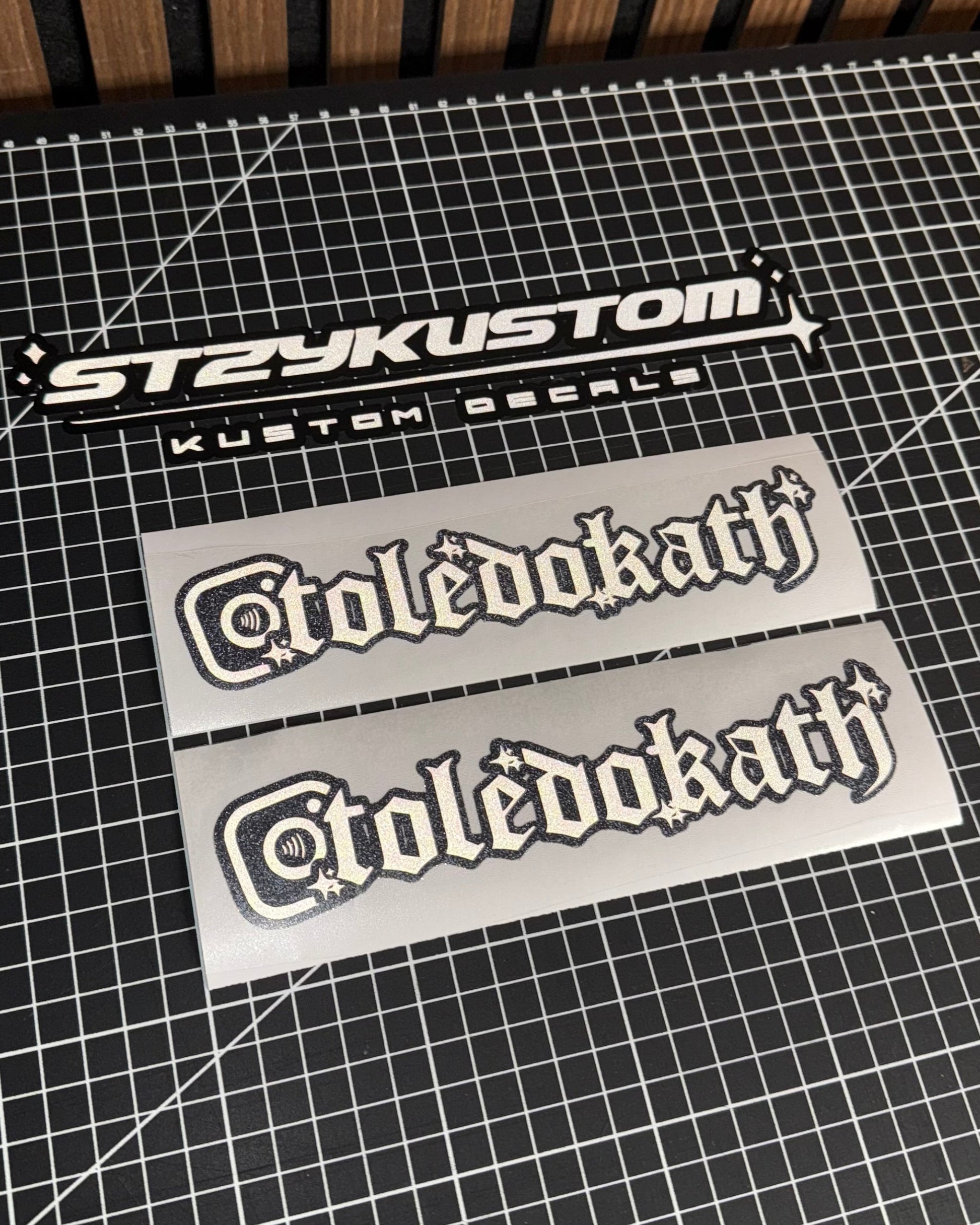 Two decals with 'Styzkustom' and 'toledokath' text on a grid surface.